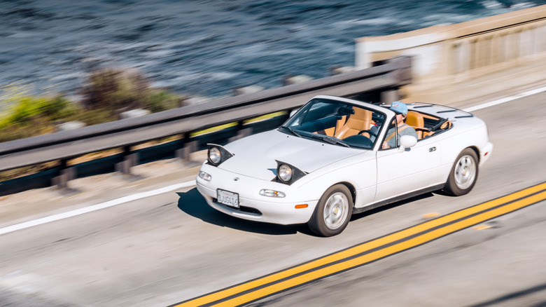 White Mazda Miata driving on coastal highway, high angle, front 3/4 view, tan interior