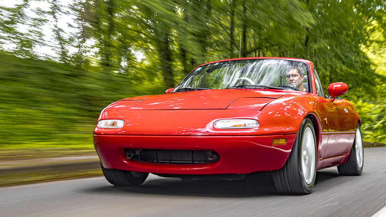 Red NA Mazda Miata on forest road, front 3/4 view