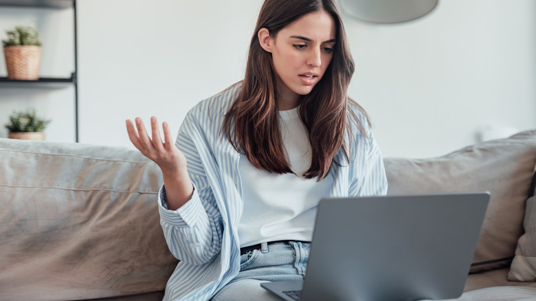 Person sitting on a couch looking upset at their laptop.