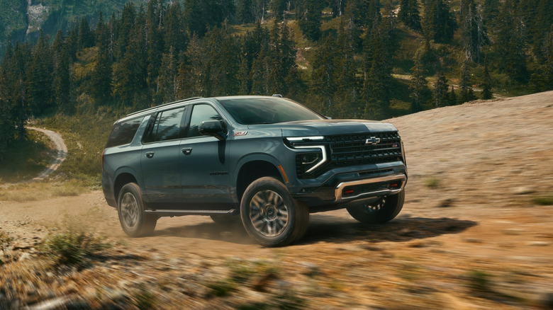 A blue 2026 Chevrolet Suburban with the Z71 package driving on a mountainous dirt road, front 3/4 view