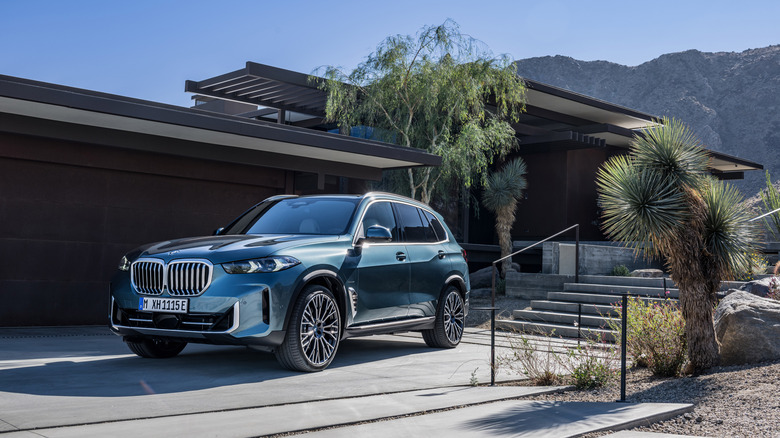 A teal 2025 BMW X5 xDrive50e outside a luxury home, front 3/4 view