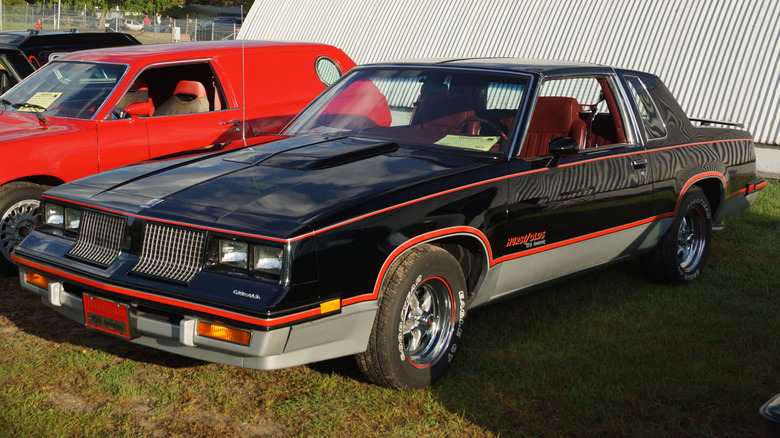 Black and silver 1983 Oldsmobile Hurst/Olds seen from left front parked near other vintage cars next to a Quonset hut