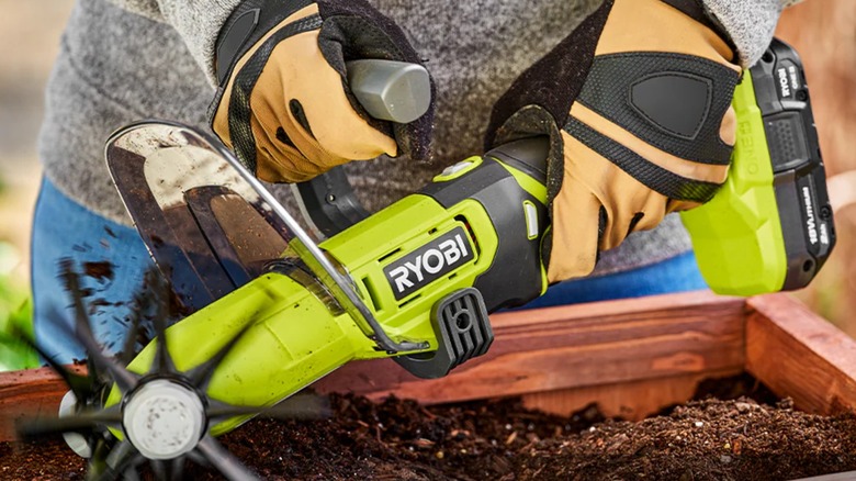 A person using a Ryobi handheld cultivator in a garden box.