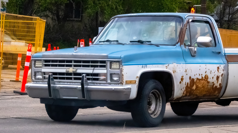 A blue-and-white Chevrolet C/K pickup truck with rusted panels on a suburban road.
