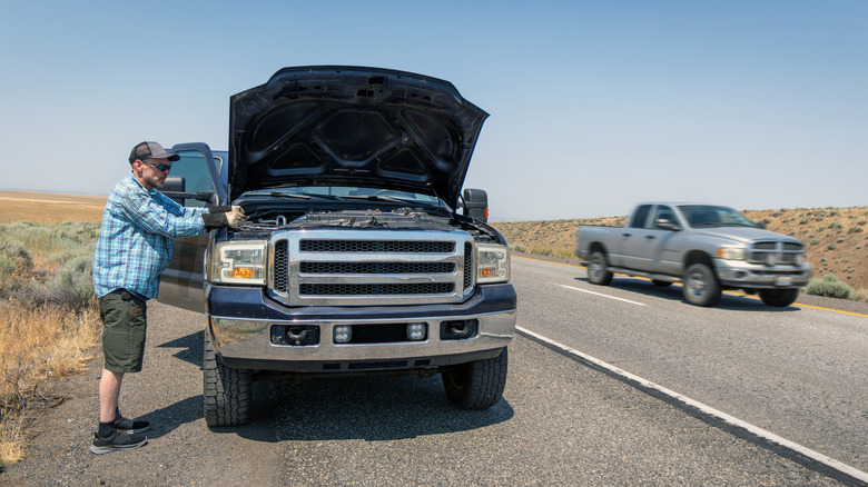 A man inspects the engine of a pickup with the hood up on side of a remote highway as another pickup drives by.