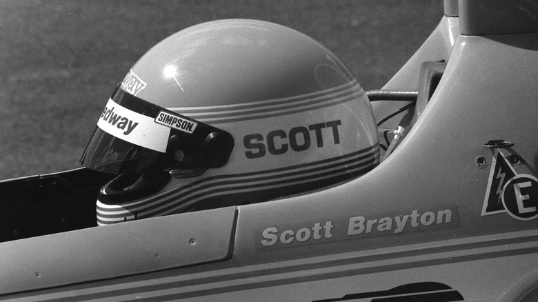 Close-up of Scott Brayton wearing racing helmet and sitting in IndyCar, left-side view, black & white