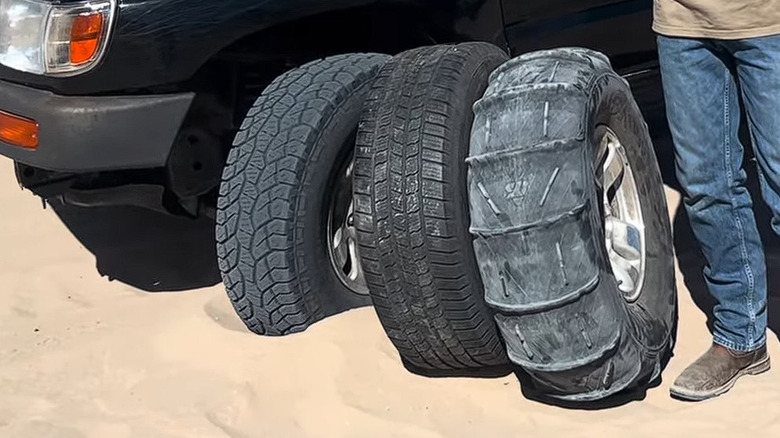An old, black Toyota SUV parked on sand, with two different typoes of tires visible next to the regular all-terrains its already wearing.