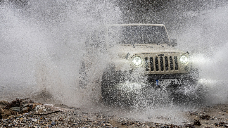 A Jeep Wrangler driving through mud and water, kicking up a shower, while off-roading.