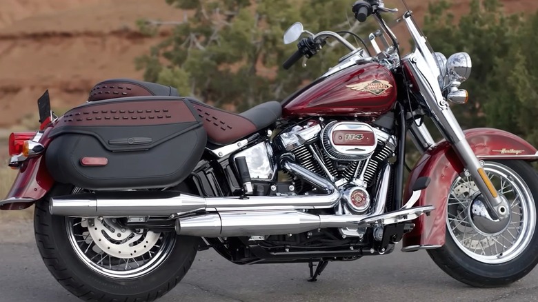 A classic Harley-Davidson Heritage cruiser in deep red with studded saddlebags and a chrome-heavy V-twin, parked outdoors against a rocky backdrop.