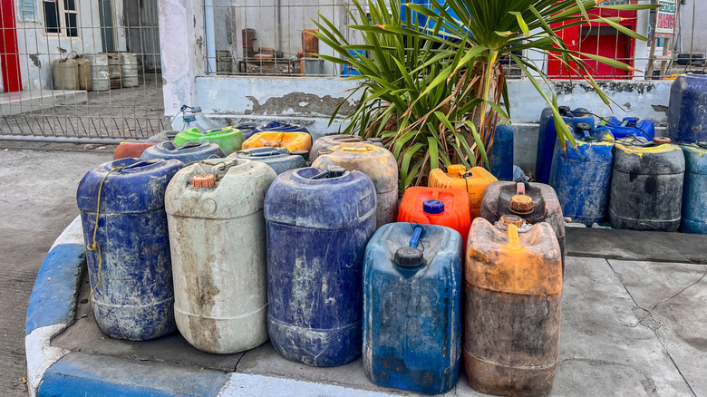 A row of used, worn-out, and old jerry cans spread across a public sidewalk near a palm tree.