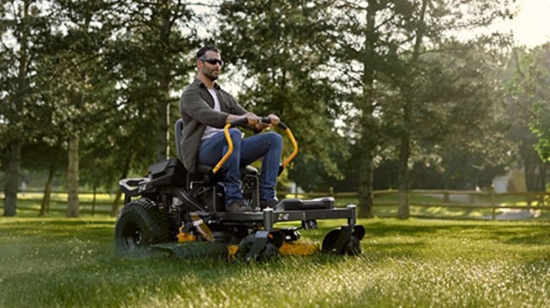 Person driving a Cub Cadet Z series lawn mower on grass with a fence and trees in the background