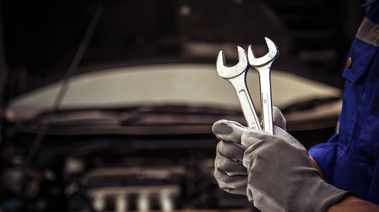 A pair of hands holding two wrenches in front of the open hood of a car
