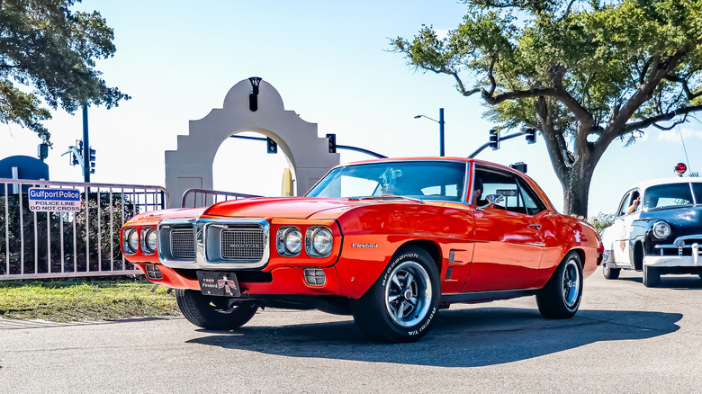 A red 1969 Pontiac Firebird rolling down a road with trees in the background