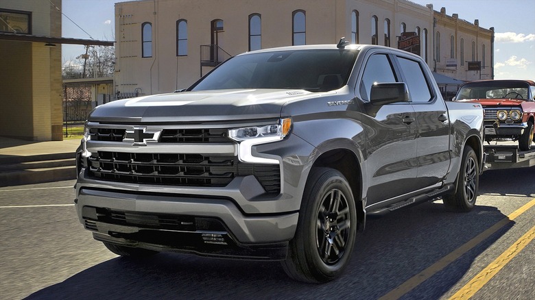 A 2026 Chevrolet Silverado 1500 RST in Sterling Gray Metallic towing a bright red vintage car on a trailer behind it in a small-town's main street on a clear day.