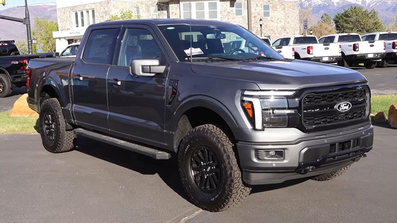 A 2026 dark gray F-150 Raptor with rugged tires is parked in a dealership lot, with rows of other trucks and mountains in its background.
