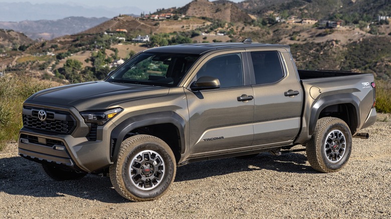 A 2025 Toyota Tacoma TRD Off-Road in Bronze Oxide with black wheels parked on a rocky dirt hillside, surrounded by dry terrains and distant homes under a clear sky.