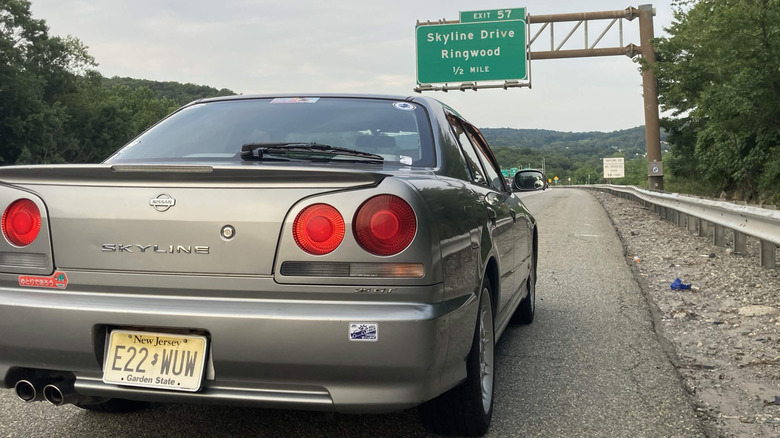 2000 Nissan Skyline R34 parked on side of highway with exit sign reading, "Skyline Rive / Ringwood" in background, rear-right close-up view