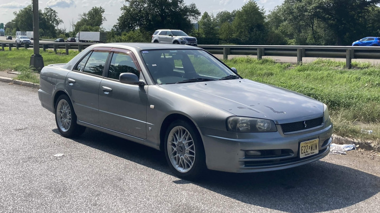 2000 Nissan Skyline R34 sedan parked in lot of wooded area with highway in background, front-right 3/4 view