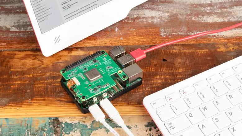 Image of Raspberry Pi AI HAT+ attached to a Raspberry Pi with several cables attached. In the background is a wooden table and the edge of a monitor and keyboard.