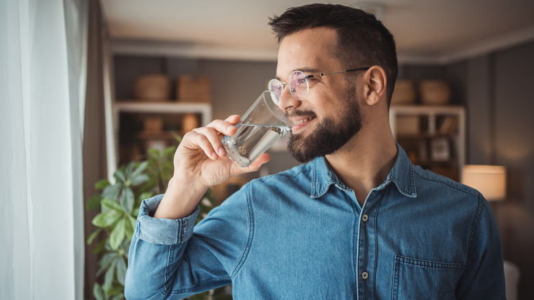 Man in blue shirt smiling and standing in home about to sip from a glass of water