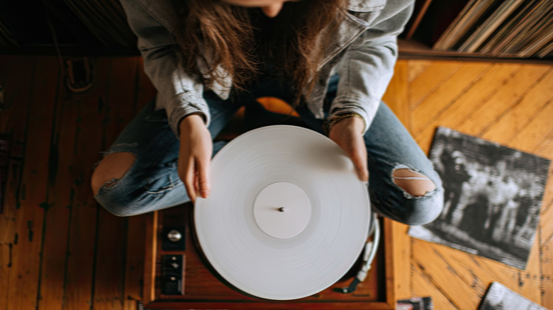 A person sitting cross-legged holding a white vinyl record over a turntable.
