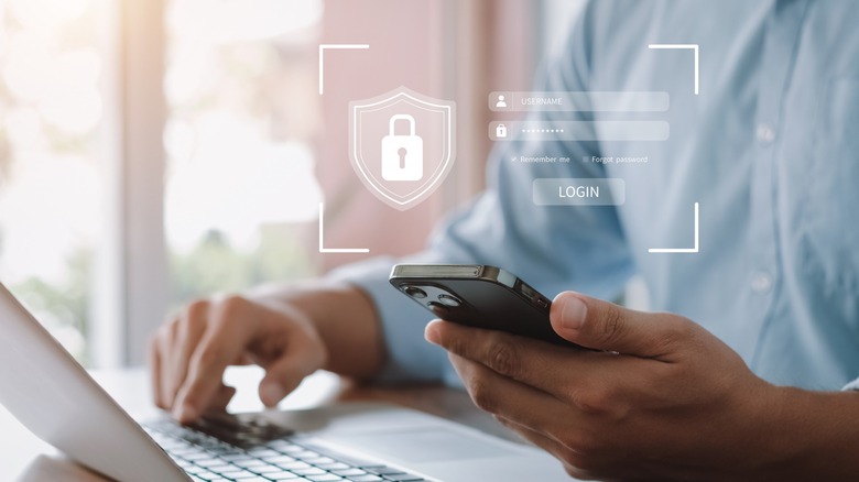 man holding cellphone in one hand while typing on a laptop with a lock icon and login screen