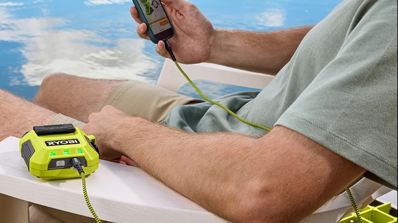 A person sitting by the water, charging their phone with the Ryobi USB Lithium 3-Port Charger and Power Source.