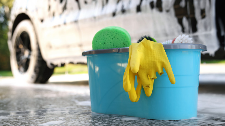 A large blue bucket with a sponge, scrub brush and yellow gloves draped over it sitting in front of a soapy vehicle