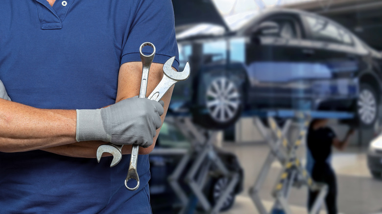 A mechanic with crossed arms holding wrenches with a blue sedan on a lift in the background