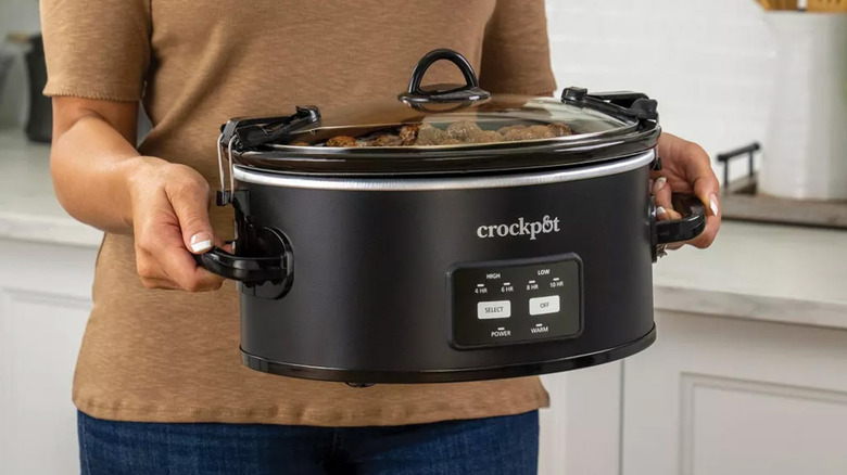 A woman carrying a Crockpot across a kitchen.