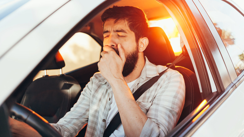 Tired driver yawning at the wheel with the scene illuminated by a setting sun.