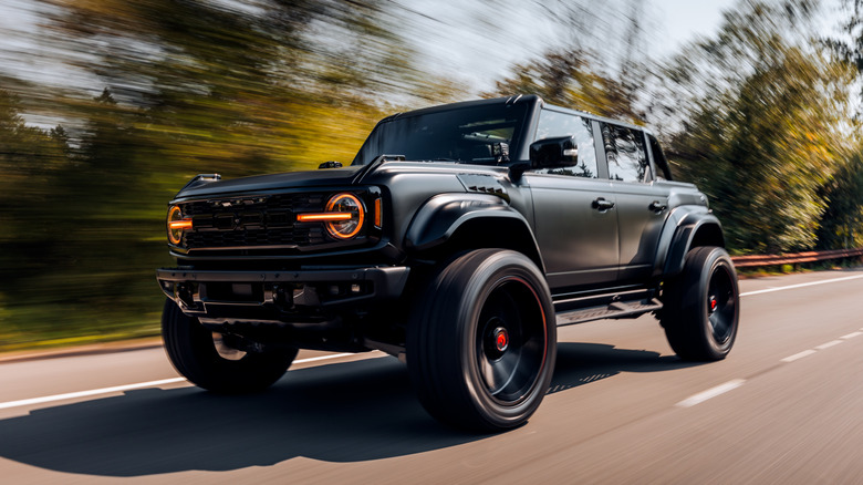 A current-generation black Ford Bronco Raptor driving down a wooded road at speed.