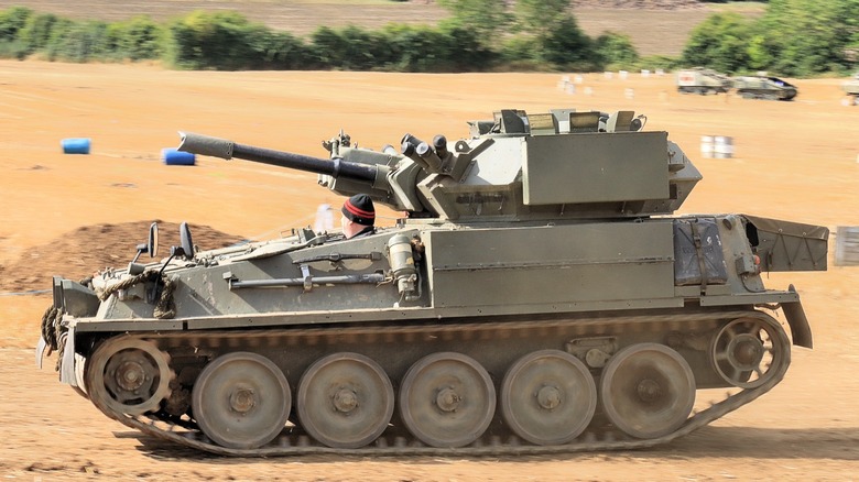 An FV101 Scorpion moving along a dirt track with its driver's head peeking out of the hatch