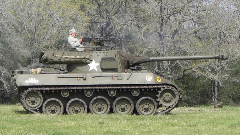A man holds an M2 Browning machine gun atop a M18 Hellcat beside a thicket of trees.