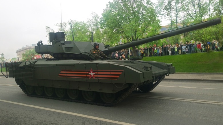 A Russian T-14 Armata MBT with its driver standing up inside its hatch as it moves along a paved road