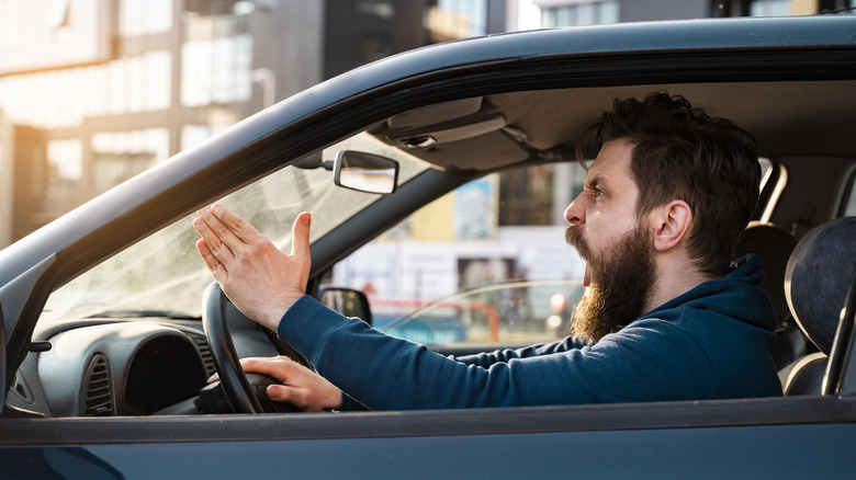 An aggressive bearded male driver sitting in a blue vehicle, yelling in frustration at something we can't see.