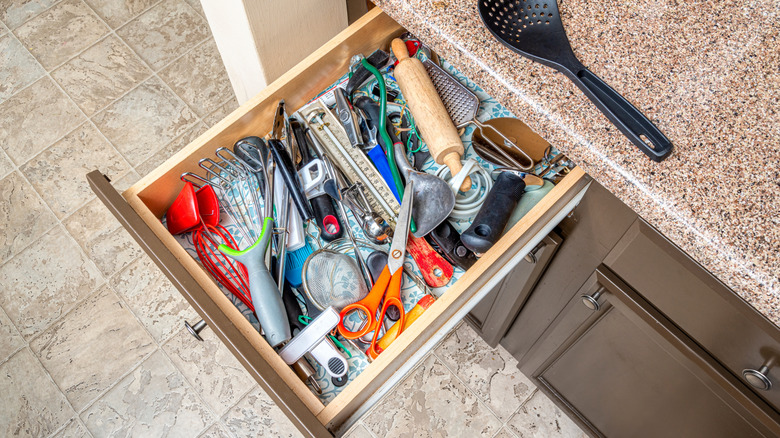 A messy kitchen drawer with a black kitchen utensil sitting on the counter above