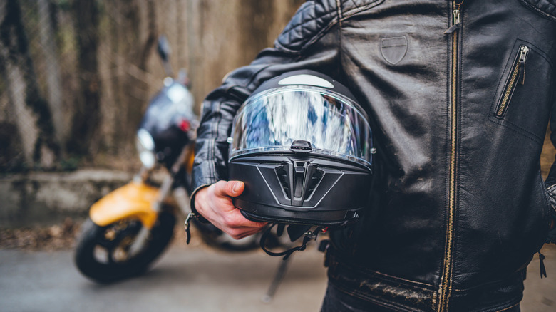 A motorcyclist in a leather riding jacket holding a helmet as they walk away from their motorbike in the background.