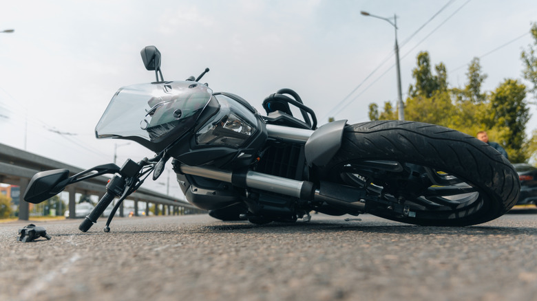 A motorcycle lying on its side, presumably after an accident on a paved concrete road in the sunlight.