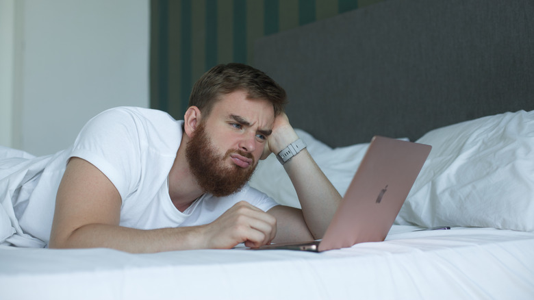 An annoyed man lying in bed with a Macbook