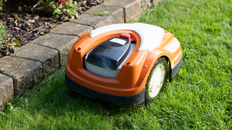 An orange, white, and black robotic lawnmower resting on green grass next to a stone garden border