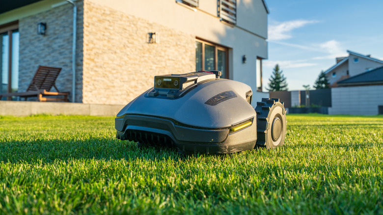 A dark gray robotic lawnmower sitting on a neatly trimmed green lawn, with a modern, two-story house in the background under a blue sky