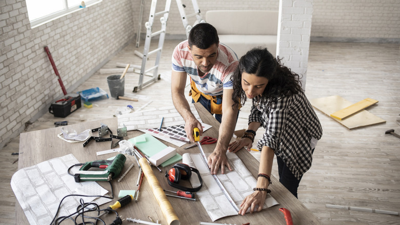A couple measuring wall covering on a table cluttered with tools in a room under construction