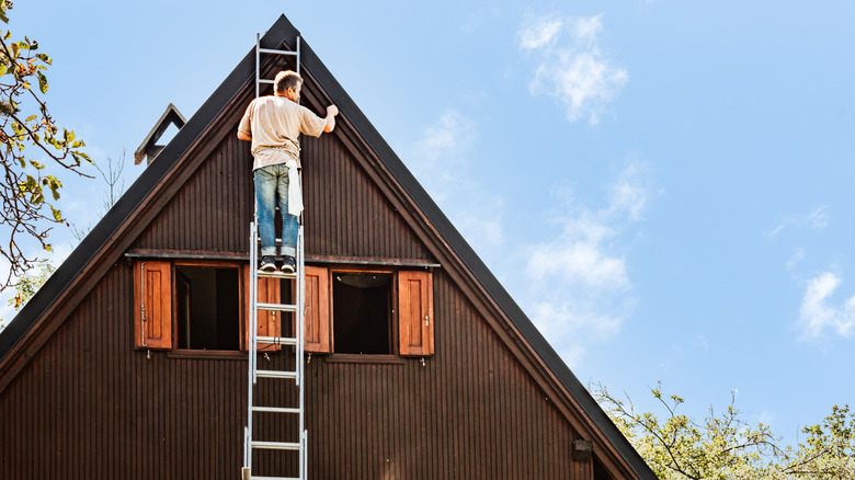 A man on a tall ladder painting a brown house