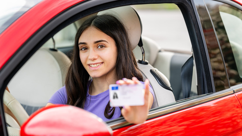 A smiling young woman sitting behind the wheel of a red vehicle and proudly holding up her driver's license