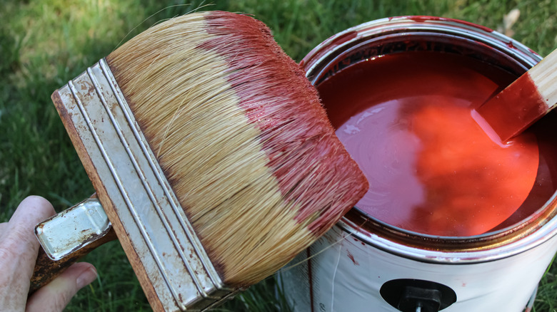 Close-up of hand holding a wide paintbrush with red paint on the tip near a gallon of red paint with a stirring stick in it