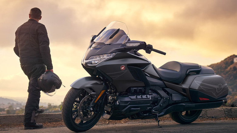 A rider in a leather jacket looks off into the sunset as his black Honda Gold Wing from 2026 is parked next to him on a twisting mountain road.