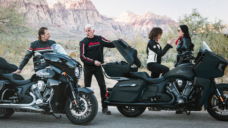Four people stand around two Indian touring motorcycles, both black, one matte and one glossy at the side of a road.