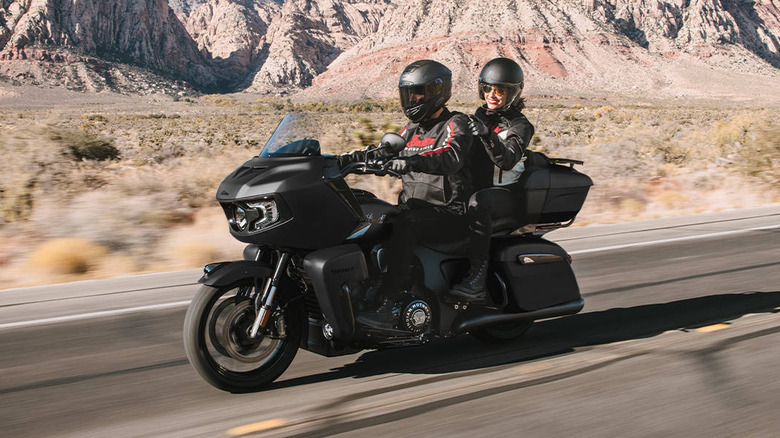 Two riders, a man and a woman, in full gear with helmets on a 2026 Indian Pursuit Dark Horse motorbike, moving at speed down a sandy highway.