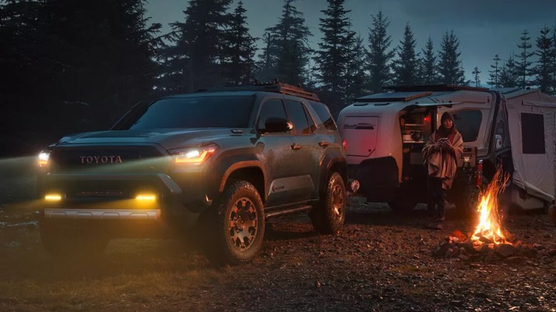 A green Toyota 4Runner with its lights on towing a camper, parked next to a campfire